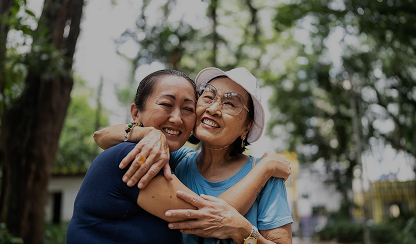 Two older Asian women smiling and hugging each other warmly in an outdoor park setting.